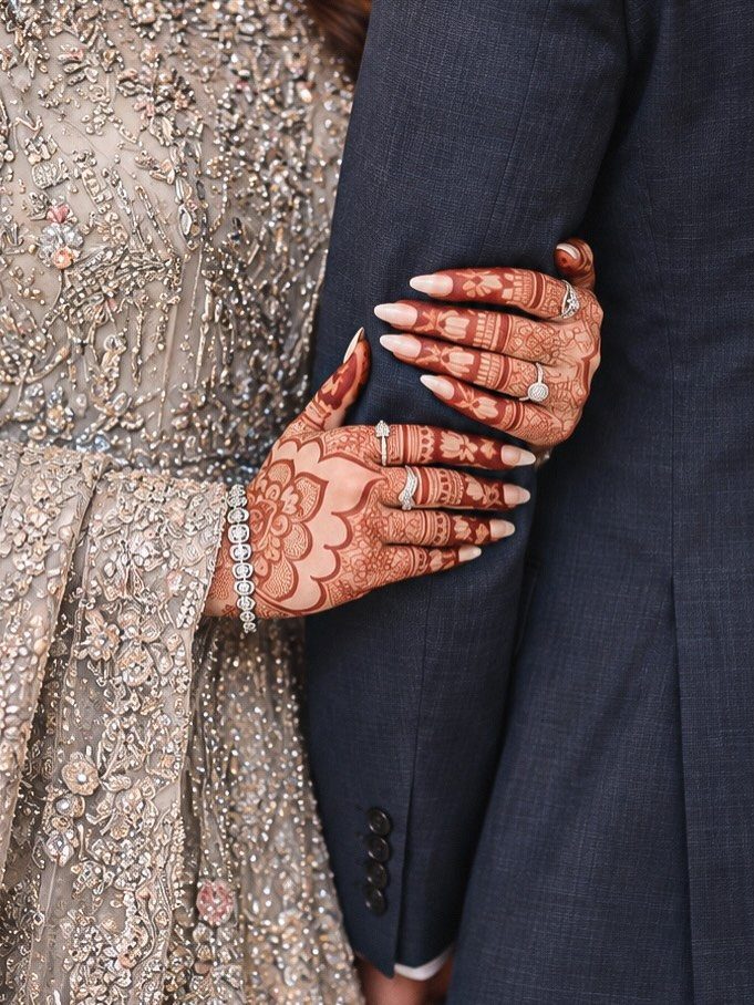 Bride's henna-decorated hands with rings resting on a groom's dark blue suit jacket