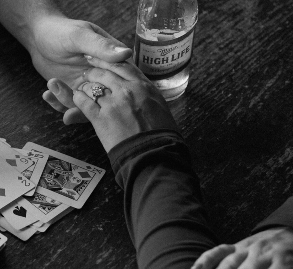 Hands with an engagement ring resting on a table with beer and cards.