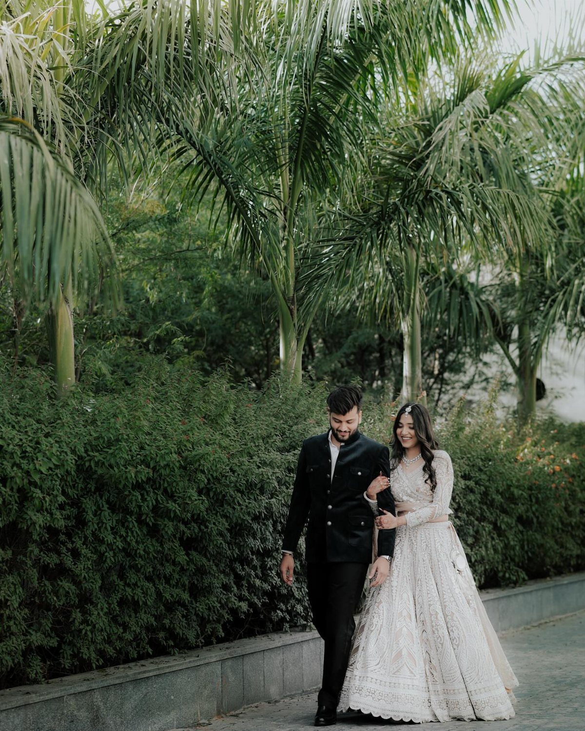 Couple in formal attire walking hand-in-hand along a path with palm trees.