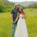 Couple posing in a green field with mountains, woman in white dress