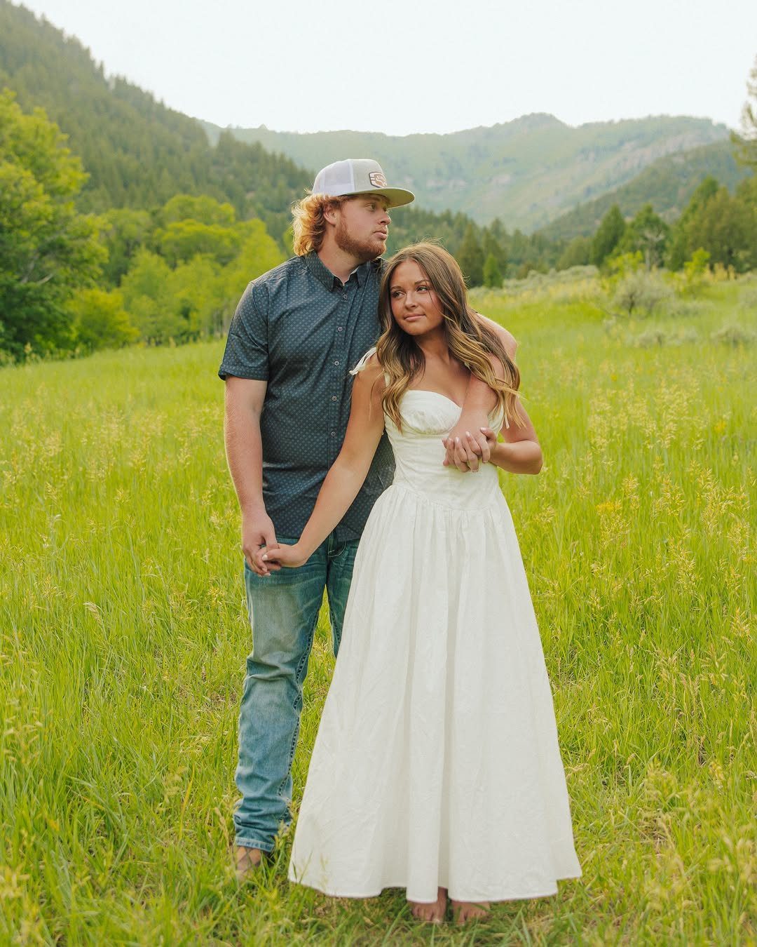 Couple posing in a green field with mountains, woman in white dress