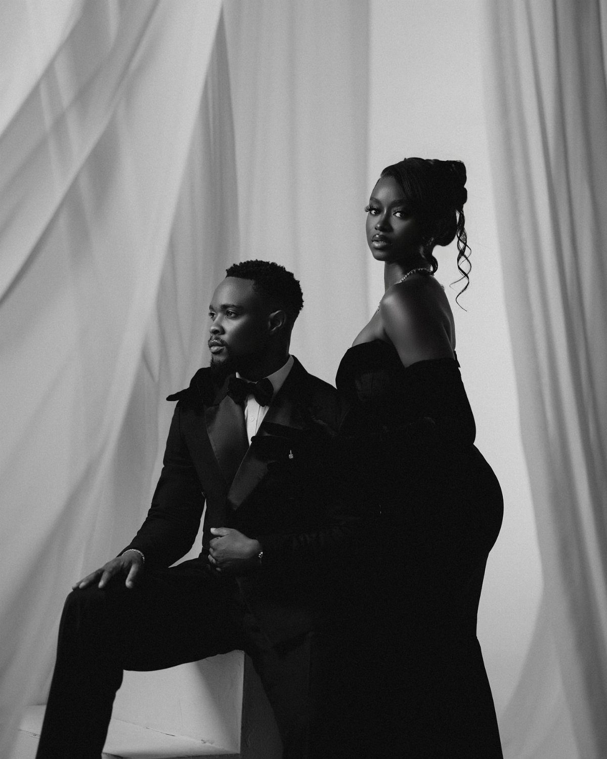 Black couple in formal attire posing against a draped white fabric backdrop.