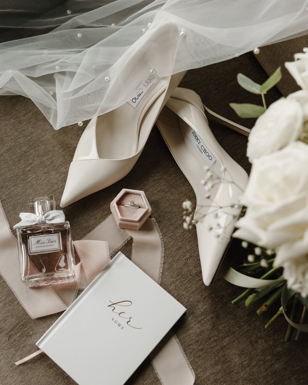 Cream wedding heels, white veil, perfume, and vow book arranged with white roses.