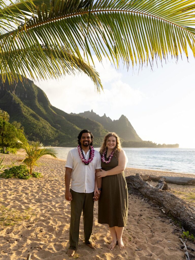 Couple wearing flower leis standing on a tropical beach with mountains in background