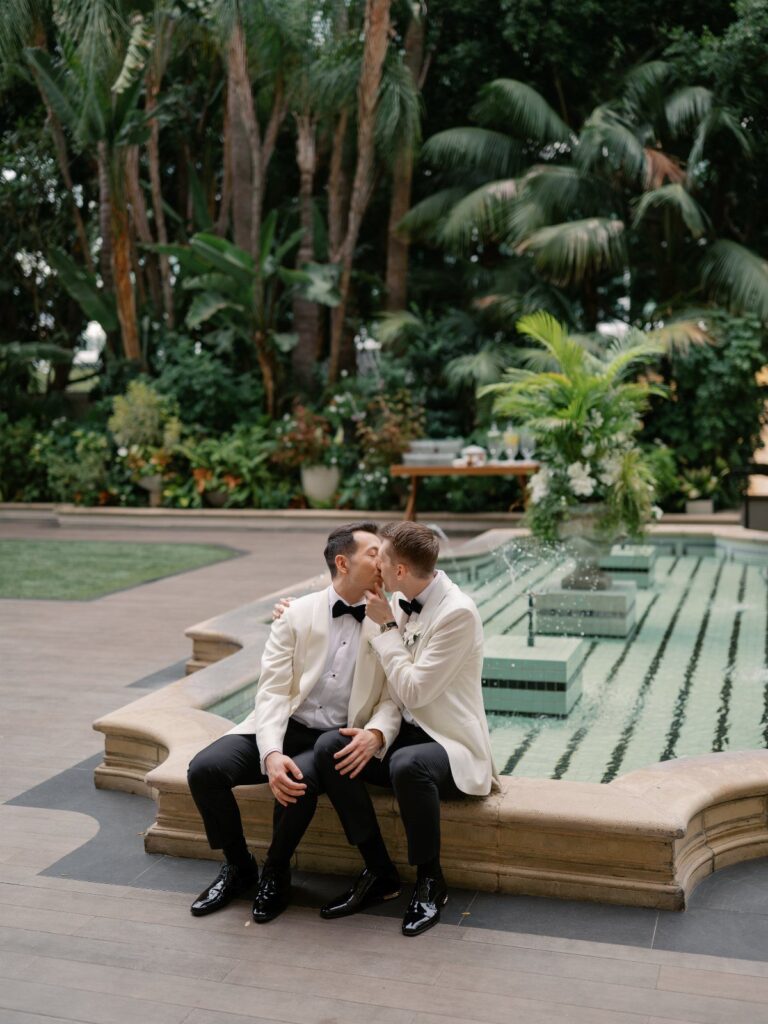 Two grooms in white tuxedos kissing by a fountain in a tropical garden