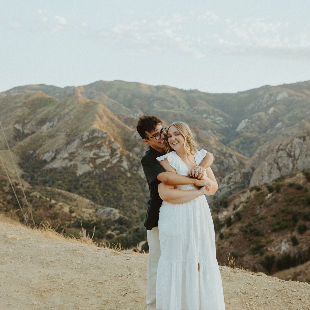 Smiling couple embracing on a dirt path with rugged mountains in the background.