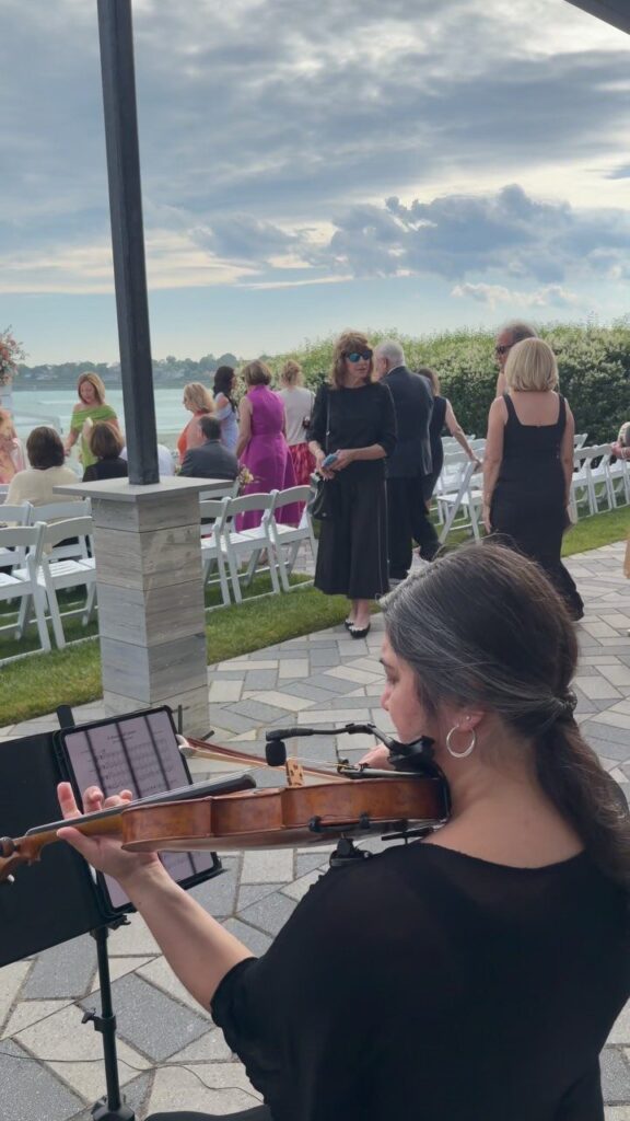 Violinist playing music for guests arriving at an outdoor wedding ceremony by the water
