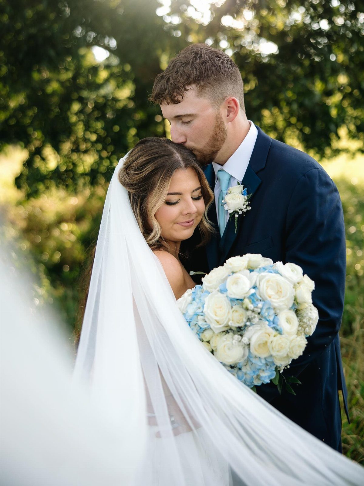 Groom kissing bride's forehead outdoors while she holds a white and blue bouquet