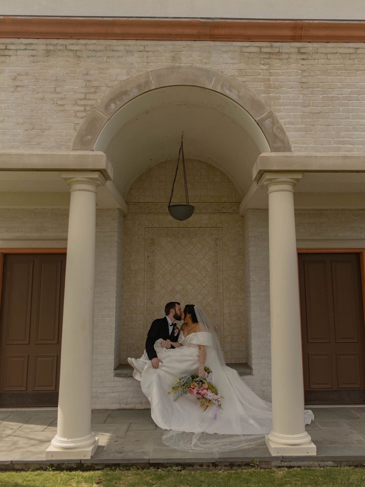 Plus-size bride in white gown kissing groom in black tuxedo in stone alcove