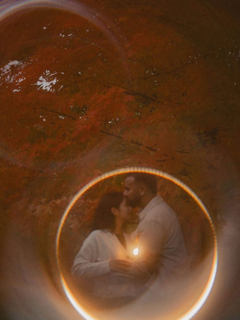 Couple kissing in a forest with autumn leaves, framed by a circular light flare.