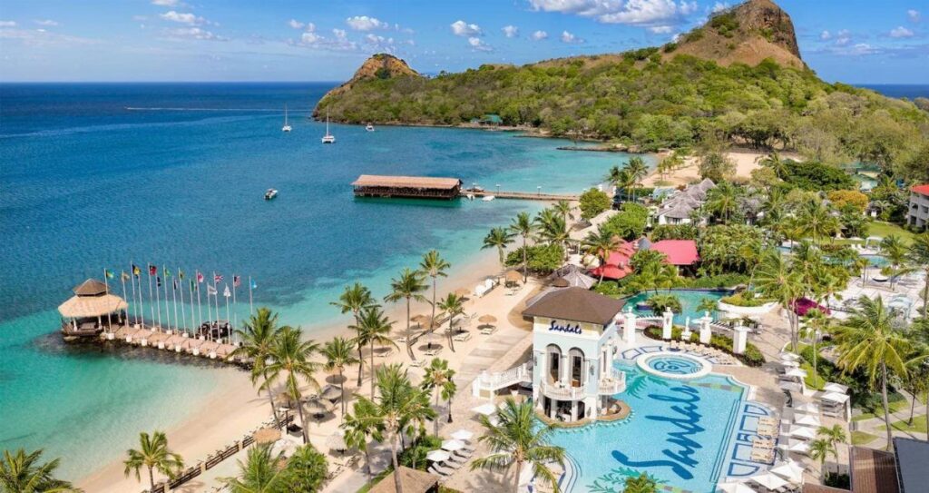 Tropical beach resort with turquoise water, swimming pool, and lush green mountains.