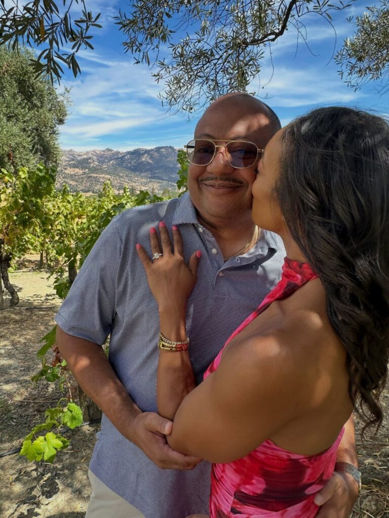 Couple embracing in a vineyard with mountains and blue sky in the background.