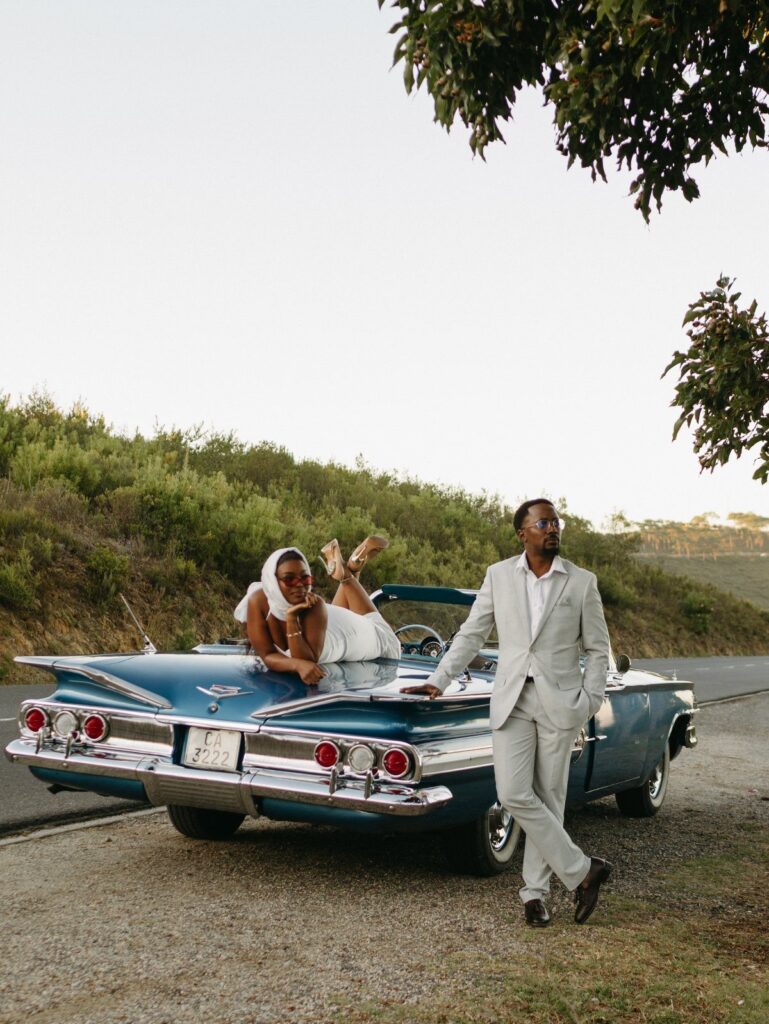 Couple posing with a blue vintage convertible car on a scenic roadside.