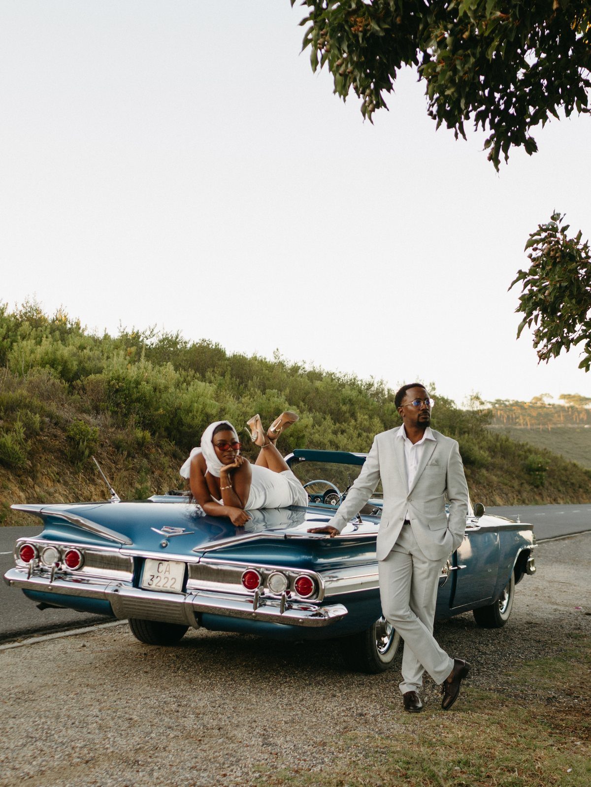 Couple posing with a blue vintage convertible car on a scenic roadside.