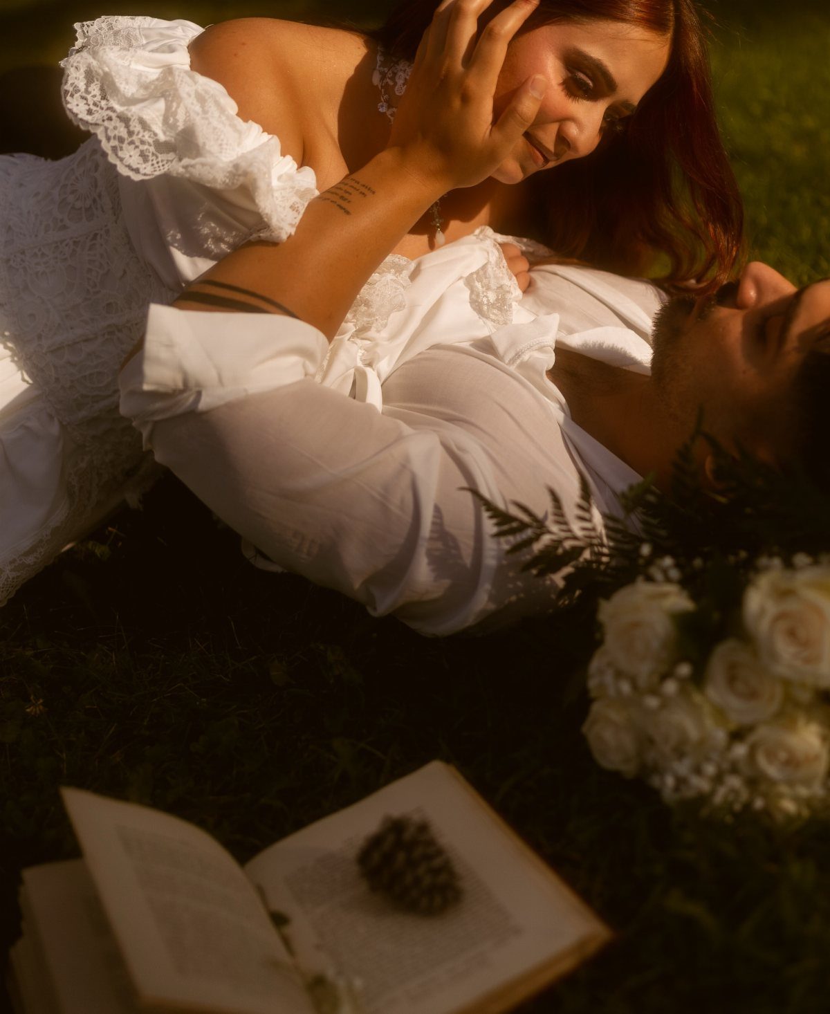 Couple in vintage white attire lying on grass with roses and an open book.