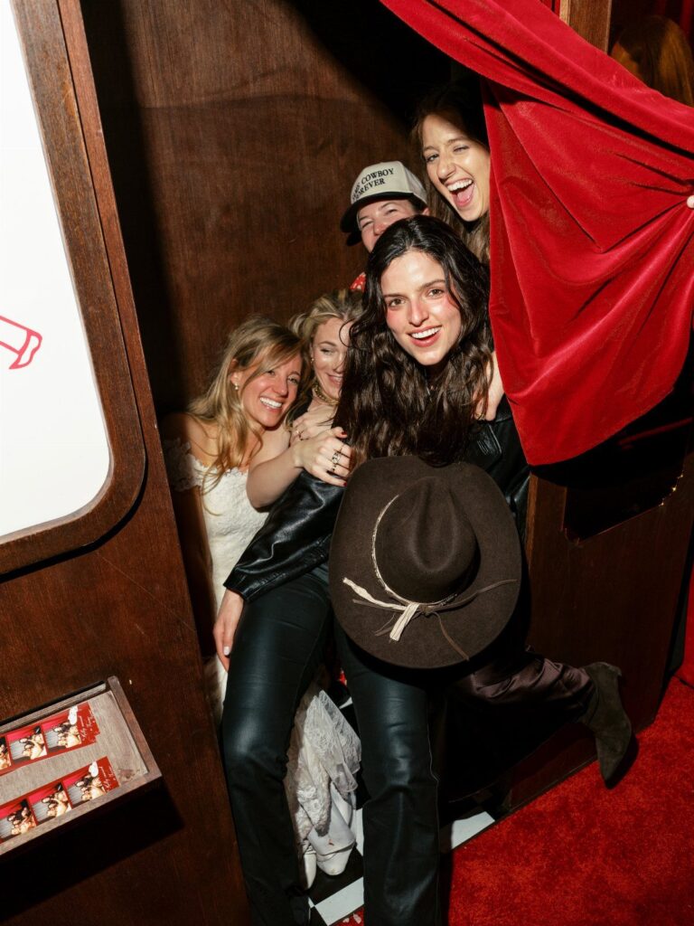 Four women posing and smiling inside a vintage photo booth with red curtains.