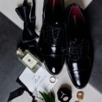 Groom's black dress shoes, bow tie, cologne, and rings arranged with a boutonniere.