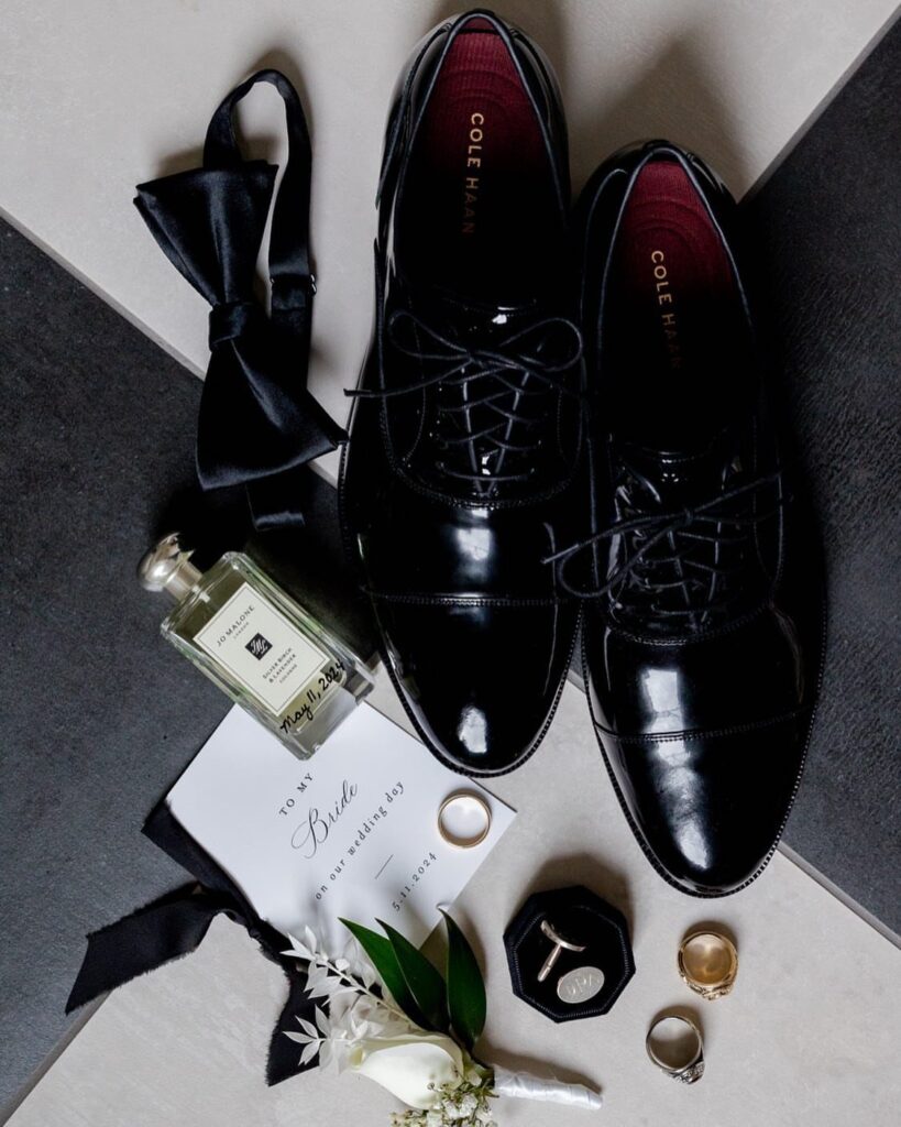 Groom's black dress shoes, bow tie, cologne, and rings arranged with a boutonniere.