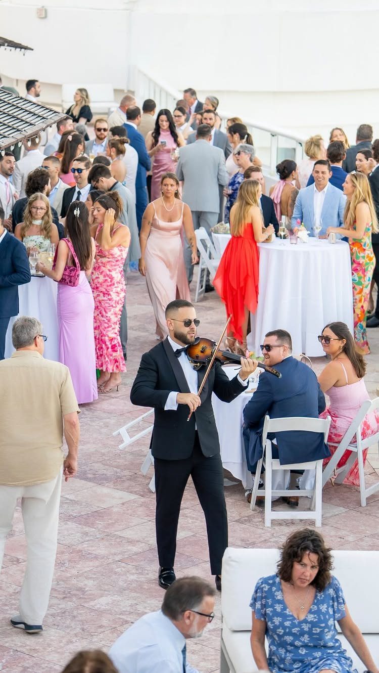 Violinist in a tuxedo playing music for guests mingling at an outdoor wedding reception.