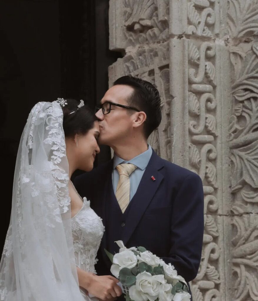 Groom in navy suit kissing the forehead of a bride in white lace dress