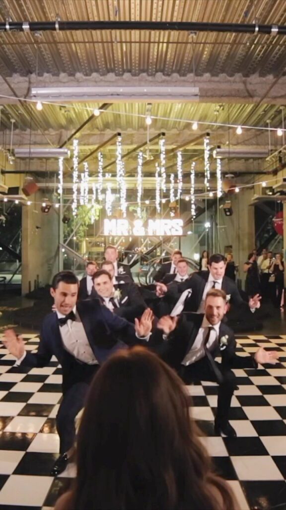 Groomsmen in tuxedos dancing on a checkered floor at a wedding reception.