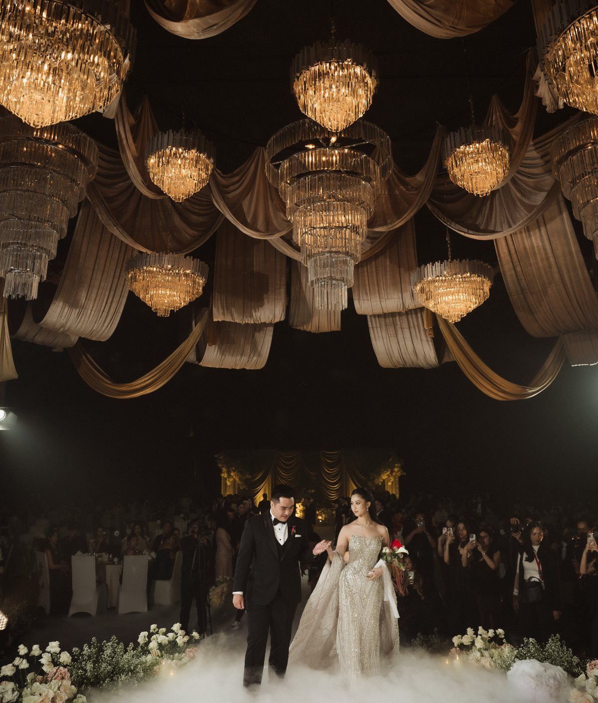 Bride and groom walking through fog under grand chandeliers at a wedding reception