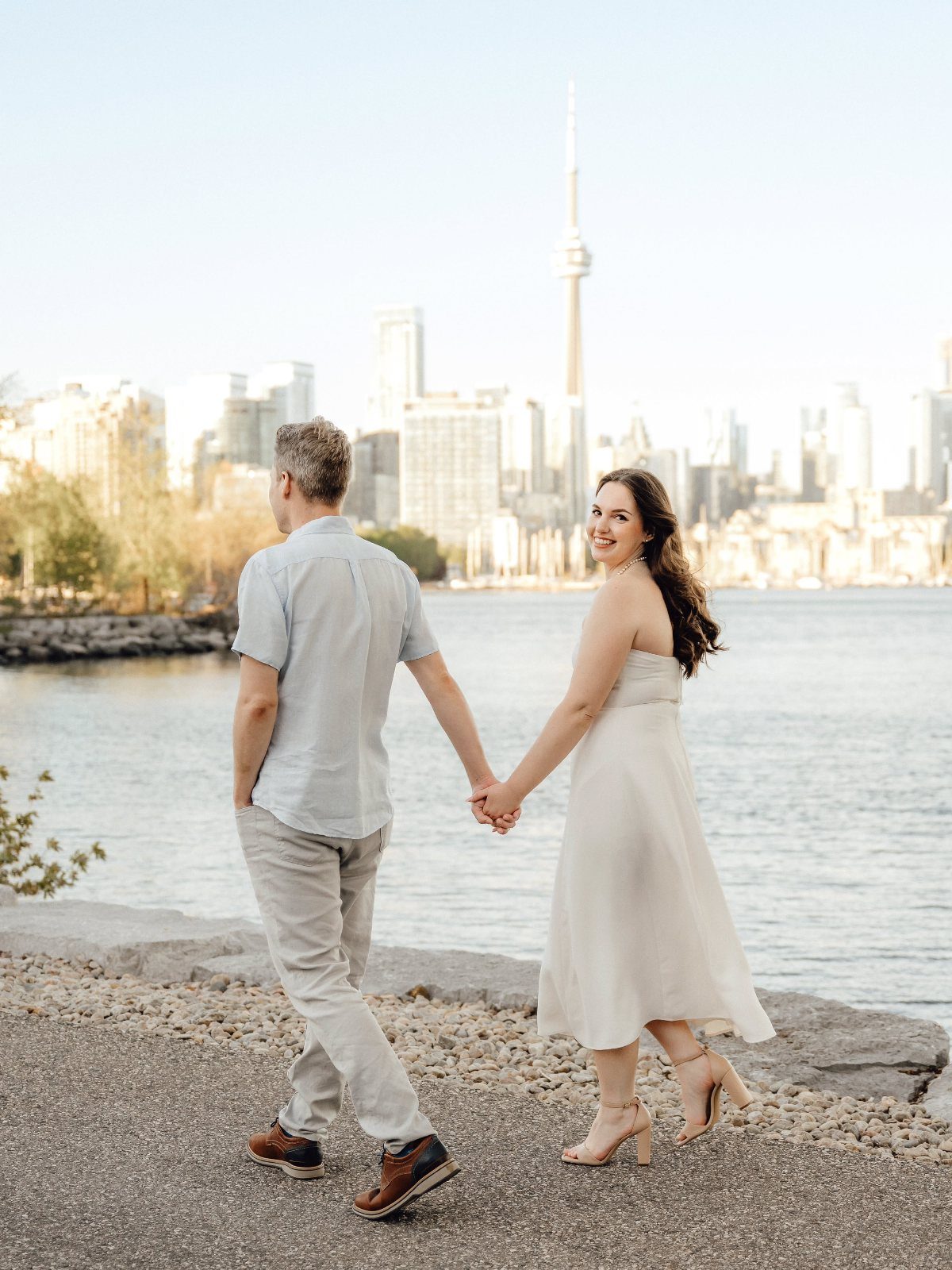 Couple in light blue and white outfits walking toward a city skyline waterfront.